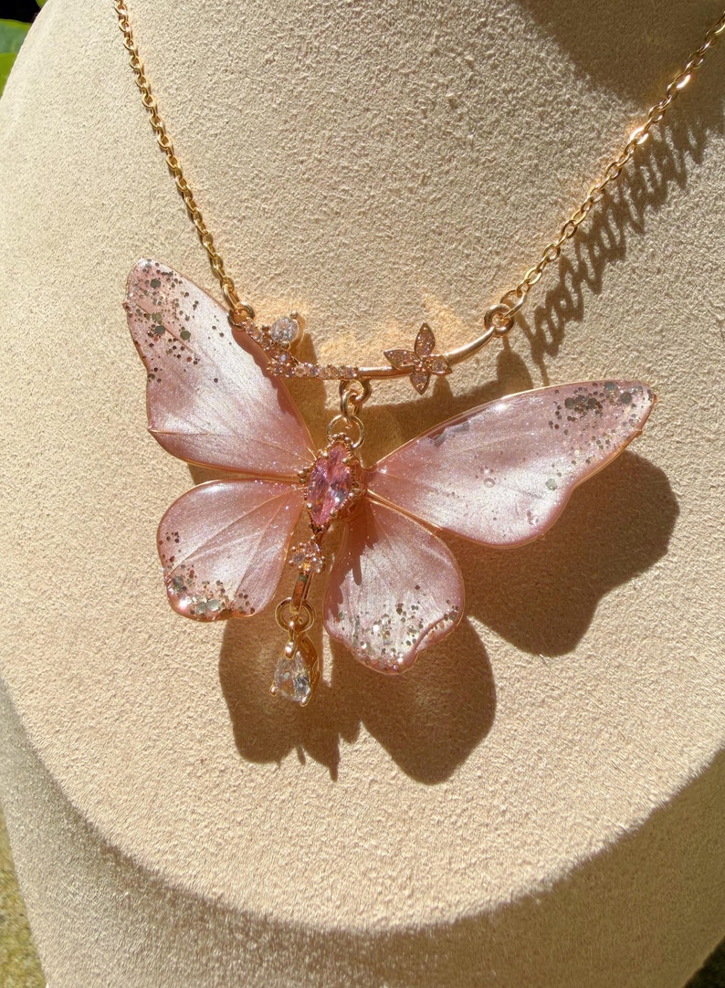 May include: A gold-tone chain necklace featuring a large, translucent pink butterfly pendant with silver glitter accents on the wing tips. The butterfly center is adorned with a marquise-cut pink crystal, and a clear teardrop crystal hangs below.