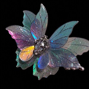 May include: A hair clip featuring translucent, iridescent butterfly wings in shades of blue, purple, and green with fine glitter accents. The center is adorned with a marquise-cut clear crystal surrounded by small, dark metallic beads.