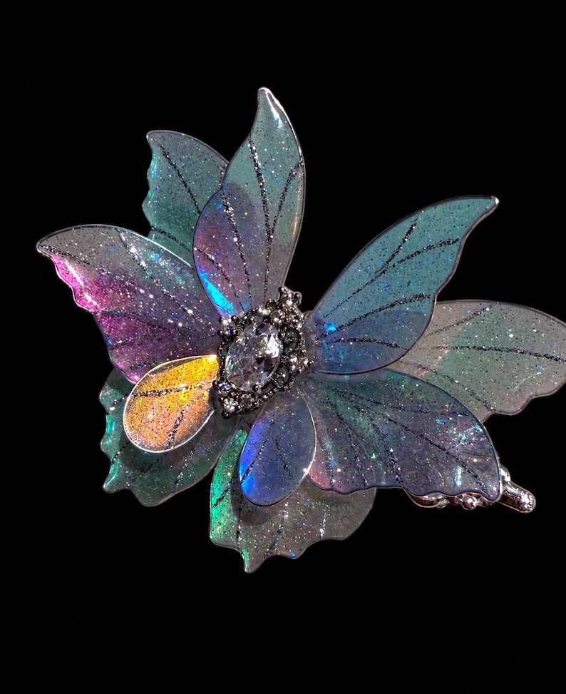May include: A hair clip featuring translucent, iridescent butterfly wings in shades of blue, purple, and green with fine glitter accents. The center is adorned with a marquise-cut clear crystal surrounded by small, dark metallic beads.