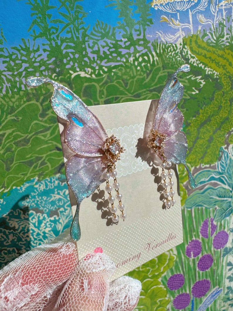 Puede incluir: Un par de pendientes de alas de mariposa iridiscentes. Las alas tienen un degradado de rosa, azul y plata, con un acento central enjoyado y cuentas de perlas colgantes. Los pendientes se muestran en una tarjeta con el texto "Blooming Versailles".