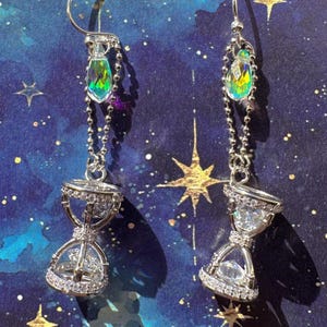 May include: A pair of silver dangle earrings with a teardrop-shaped iridescent crystal and a clear crystal hourglass charm.