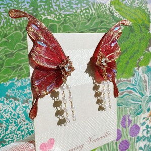 Puede incluir: Un par de pendientes de alas de mariposa rojas y doradas con detalles de perlas. Los pendientes se exhiben en una tarjeta con el texto "Blooming Versailles" y un coraz&oacute;n rosa. Las alas tienen un acabado brillante.