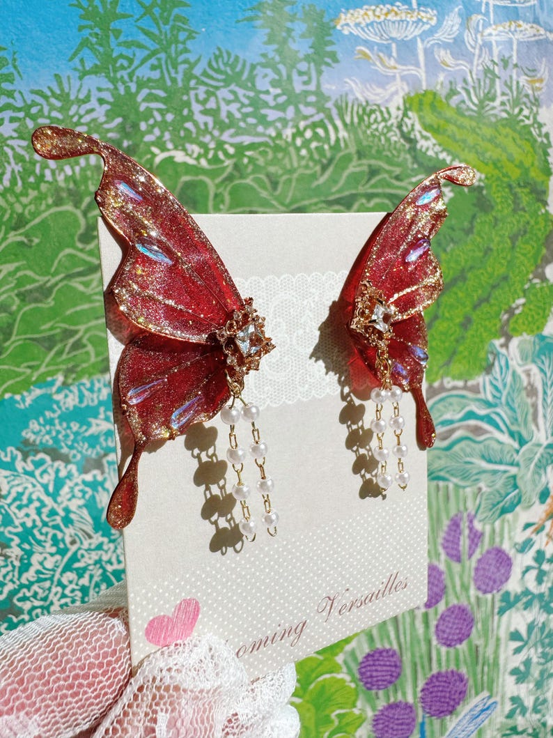 Puede incluir: Un par de pendientes de alas de mariposa rojas y doradas con detalles de perlas. Los pendientes se exhiben en una tarjeta con el texto "Blooming Versailles" y un coraz&oacute;n rosa. Las alas tienen un acabado brillante.