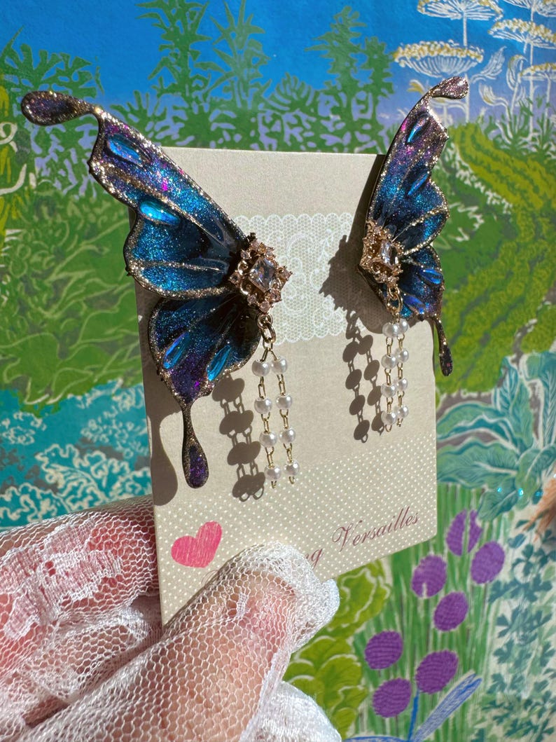 Puede incluir: Pendientes de alas de mariposa azules y moradas con detalles dorados y cadenas de perlas colgantes. Se exhiben en una tarjeta con la palabra "Versalles" y un coraz&oacute;n rosa.