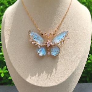May include: A gold-toned necklace featuring a butterfly pendant with translucent, iridescent blue wings. The butterfly is accented with small clear crystals along the edges and a central rectangular crystal, suspended from a delicate gold-toned chain.