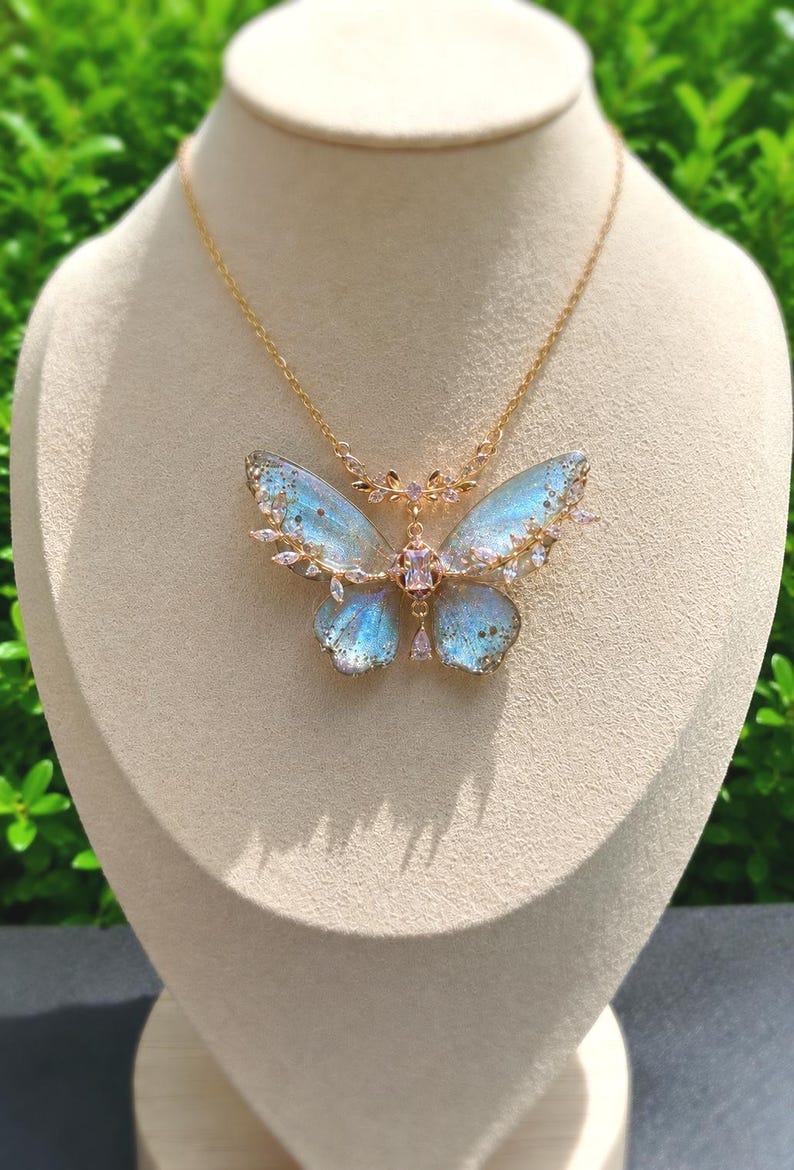 May include: A gold-toned necklace featuring a butterfly pendant with translucent, iridescent blue wings. The butterfly is accented with small clear crystals along the edges and a central rectangular crystal, suspended from a delicate gold-toned chain.