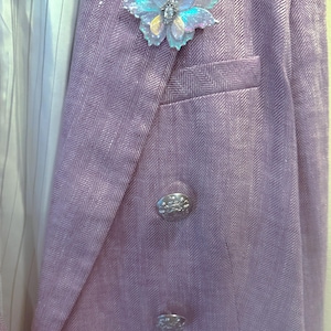 May include: A lavender herringbone blazer featuring a double-breasted front with two silver-toned embossed buttons and a welt pocket. An iridescent, translucent butterfly brooch with a central crystal accent is pinned to the left lapel.