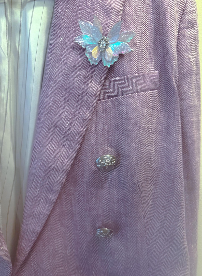 May include: A lavender herringbone blazer featuring a double-breasted front with two silver-toned embossed buttons and a welt pocket. An iridescent, translucent butterfly brooch with a central crystal accent is pinned to the left lapel.