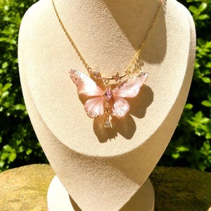 May include: A gold-tone chain necklace featuring a large, translucent pale pink butterfly pendant. The butterfly wings have subtle silver-colored flecks, a central marquise-cut pink stone, and a small, clear teardrop-shaped crystal dangling below.