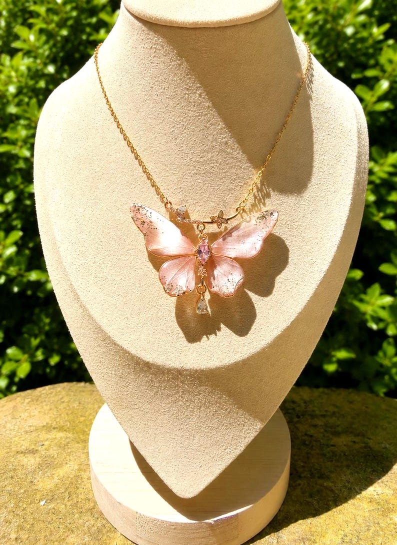 May include: A gold-tone chain necklace featuring a large, translucent pale pink butterfly pendant. The butterfly wings have subtle silver-colored flecks, a central marquise-cut pink stone, and a small, clear teardrop-shaped crystal dangling below.