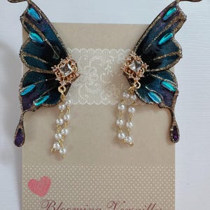 Puede incluir: Pendientes de alas de mariposa. Las alas son de color verde azulado oscuro con ribetes dorados y detalles turquesa y morados. Cada pendiente tiene un centro cuadrado dorado con cristales transparentes y una hebra de perlas blancas. El texto "Blooming Versailles" es visible.