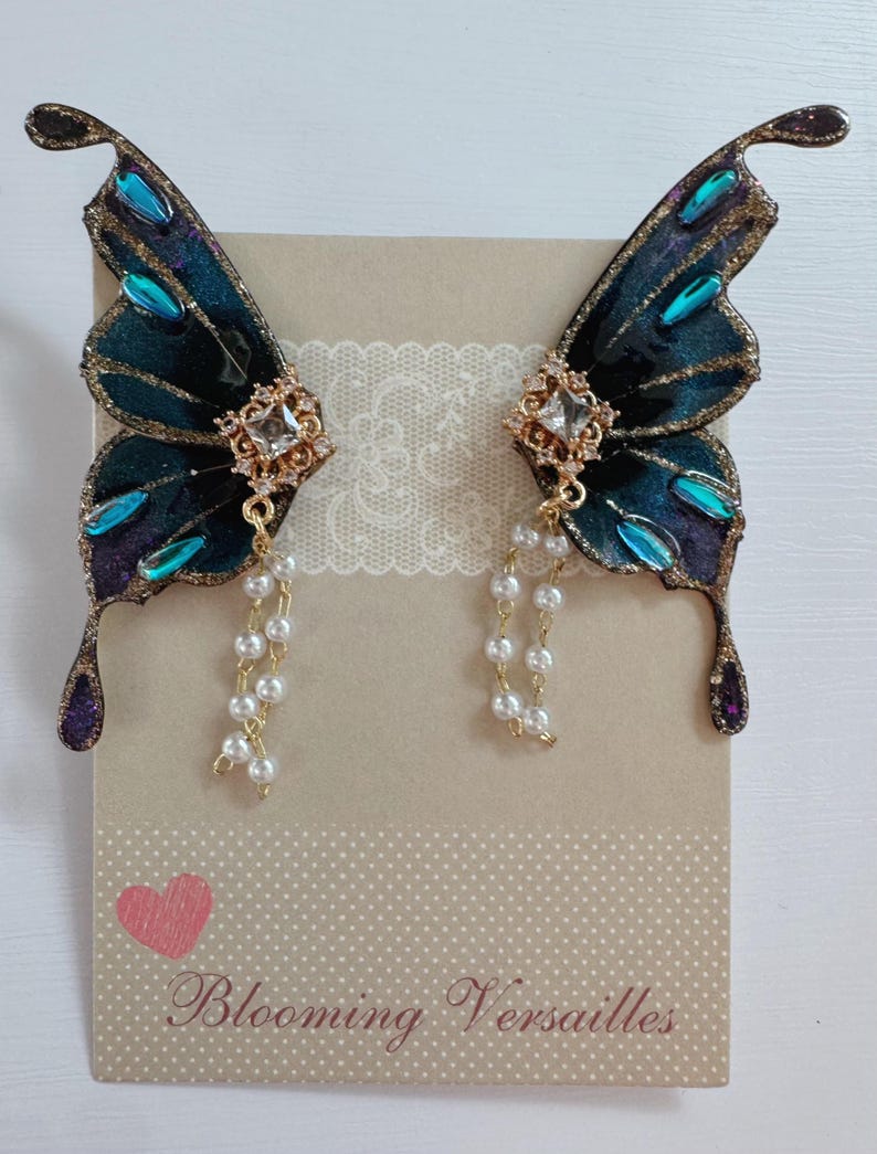 Puede incluir: Pendientes de alas de mariposa. Las alas son de color verde azulado oscuro con ribetes dorados y detalles turquesa y morados. Cada pendiente tiene un centro cuadrado dorado con cristales transparentes y una hebra de perlas blancas. El texto "Blooming Versailles" es visible.