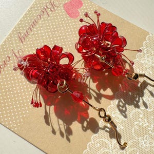 May include: A pair of red floral earrings with gold-colored accents. The earrings feature a cluster of red, translucent petals and small, dangling red beads. The earrings are displayed on a beige card with the words "Blooming Vers" and a pink heart.