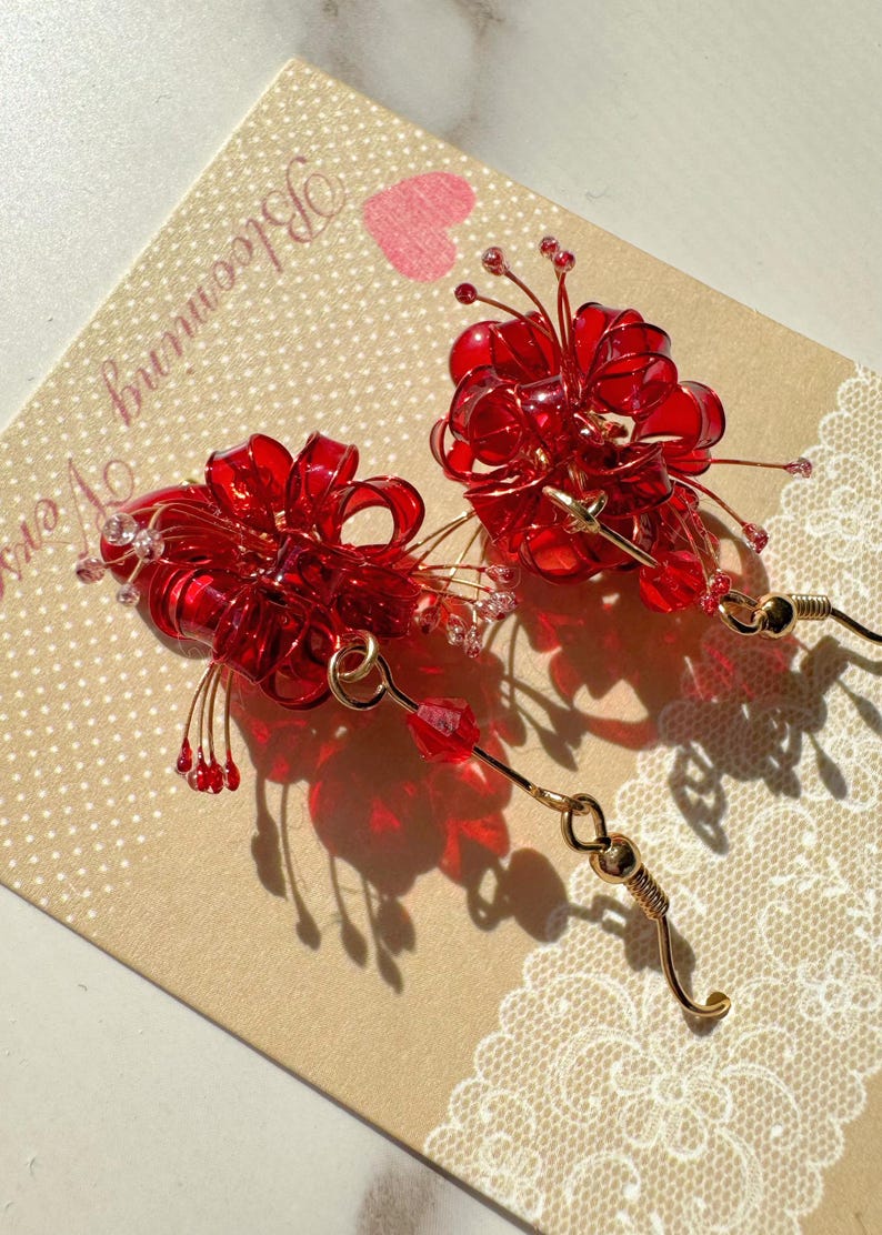 May include: A pair of red floral earrings with gold-colored accents. The earrings feature a cluster of red, translucent petals and small, dangling red beads. The earrings are displayed on a beige card with the words "Blooming Vers" and a pink heart.