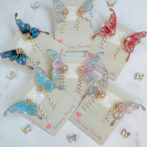 Puede incluir: Pendientes de alas de mariposa en varios colores, incluyendo azul, rosa y rojo. Cada pendiente presenta un delicado dise&ntilde;o de ala con detalles de perlas y un centro de pedrer&iacute;a. Los pendientes se exhiben en tarjetas con el texto "Blooming Versailles".