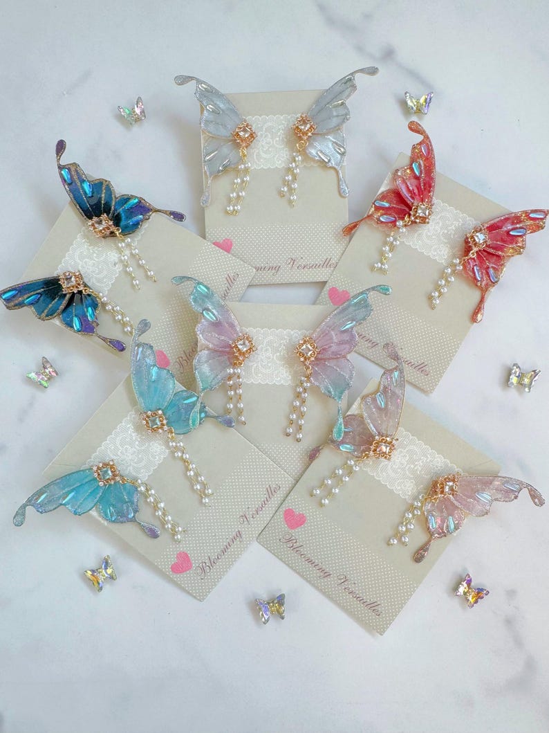 Puede incluir: Pendientes de alas de mariposa en varios colores, incluyendo azul, rosa y rojo. Cada pendiente presenta un delicado dise&ntilde;o de ala con detalles de perlas y un centro de pedrer&iacute;a. Los pendientes se exhiben en tarjetas con el texto "Blooming Versailles".