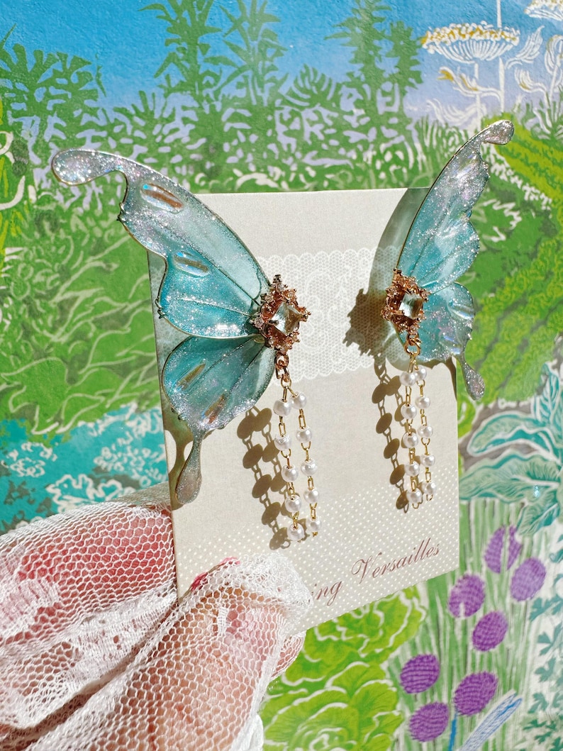Puede incluir: Un par de pendientes de alas de mariposa azul iridiscente con detalles de purpurina plateada. Cada pendiente presenta un centro de filigrana dorada con hilos colgantes de perlas de imitaci&oacute;n. Los pendientes se muestran en una tarjeta color crema con el texto "Versailles".