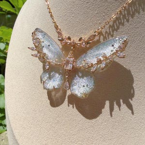May include: A rose gold necklace featuring a large butterfly pendant with translucent, glitter-infused wings. The butterfly is accented with clear, marquise-cut crystals along the edges and a central rectangular stone, suspended from a delicate chain.