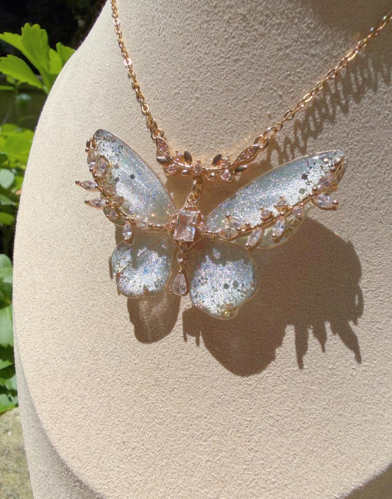 May include: A rose gold necklace featuring a large butterfly pendant with translucent, glitter-infused wings. The butterfly is accented with clear, marquise-cut crystals along the edges and a central rectangular stone, suspended from a delicate chain.