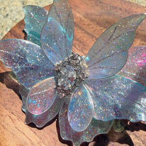 May include: A butterfly-shaped hair accessory featuring translucent, iridescent wings with a fine glitter finish. The center is adorned with a cluster of clear, faceted crystals, including a prominent oval-shaped stone surrounded by smaller round stones.