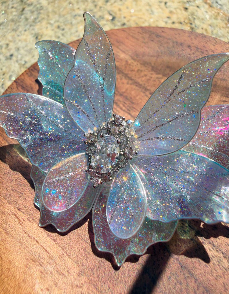 May include: A butterfly-shaped hair accessory featuring translucent, iridescent wings with a fine glitter finish. The center is adorned with a cluster of clear, faceted crystals, including a prominent oval-shaped stone surrounded by smaller round stones.