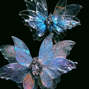 May include: Two butterfly-shaped hair clips featuring translucent, iridescent wings with fine glitter accents. Each clip has a central metallic body adorned with a clear, faceted gemstone. The wings display shifting blue, purple, and pink hues.