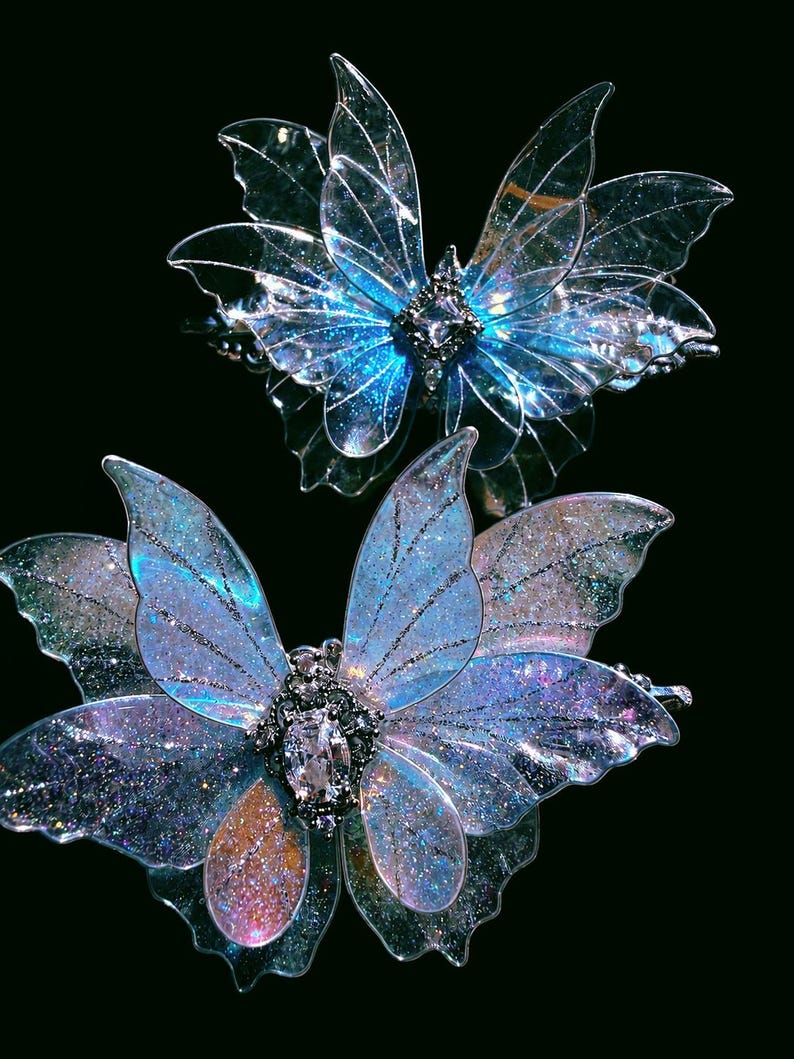 May include: Two butterfly-shaped hair clips featuring translucent, iridescent wings with fine glitter accents. Each clip has a central metallic body adorned with a clear, faceted gemstone. The wings display shifting blue, purple, and pink hues.