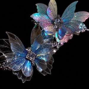 May include: Two hair clips featuring translucent, iridescent butterfly wings in shades of blue, purple, and pink. Each butterfly has a central square-cut clear crystal accent surrounded by small silver-toned metal detailing and glitter.