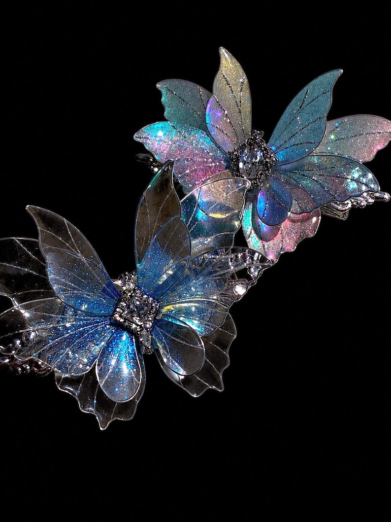 May include: Two hair clips featuring translucent, iridescent butterfly wings in shades of blue, purple, and pink. Each butterfly has a central square-cut clear crystal accent surrounded by small silver-toned metal detailing and glitter.