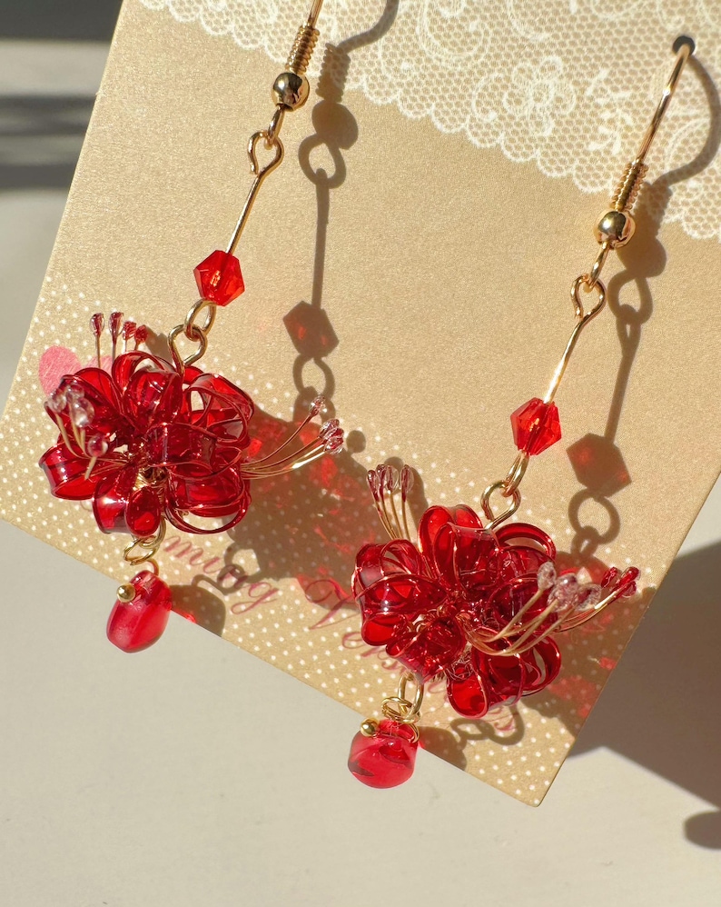 May include: A pair of red floral earrings with gold-colored hooks. Each earring features a wire-formed flower with red beads and a dangling red gem. The earrings are displayed on a light brown card.