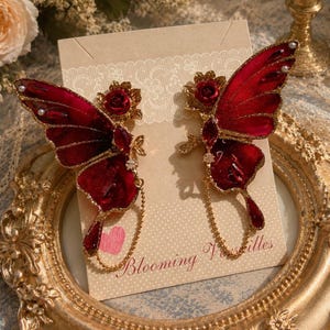 May include: A pair of ornate red butterfly earrings with gold accents, featuring a rose detail and dangling chain. The earrings are displayed on a decorative gold frame with the text "Blooming Versailles" on a card.