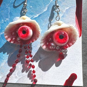 May include: A pair of unique earrings featuring a white seashell design, each holding a red eyeball with a black pupil. The earrings are adorned with red beads and silver hooks, creating a striking and unusual accessory.
