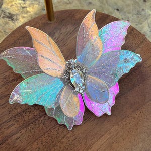 May include: A butterfly-shaped hair accessory featuring iridescent, translucent wings with silver glitter accents. The center is adorned with a clear, oval-shaped faceted crystal surrounded by small, clear rhinestones set in a silver-toned metal base.