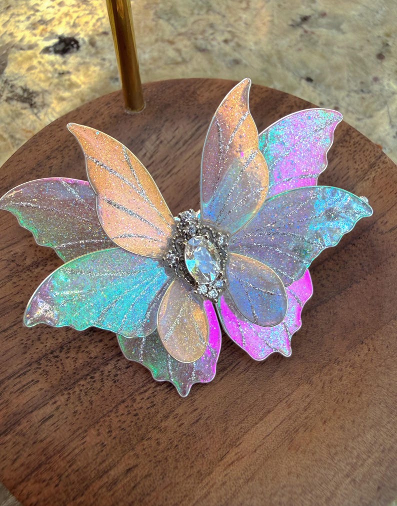 May include: A butterfly-shaped hair accessory featuring iridescent, translucent wings with silver glitter accents. The center is adorned with a clear, oval-shaped faceted crystal surrounded by small, clear rhinestones set in a silver-toned metal base.