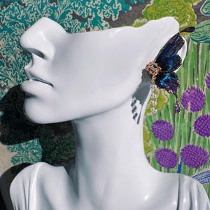 Puede incluir: Un pendiente de oreja con forma de mariposa en tonos azules y morados, adornado con detalles dorados y colgantes de perlas y gemas moradas. El pendiente se exhibe en una cabeza de maniqu&iacute; blanca sobre un fondo floral colorido.