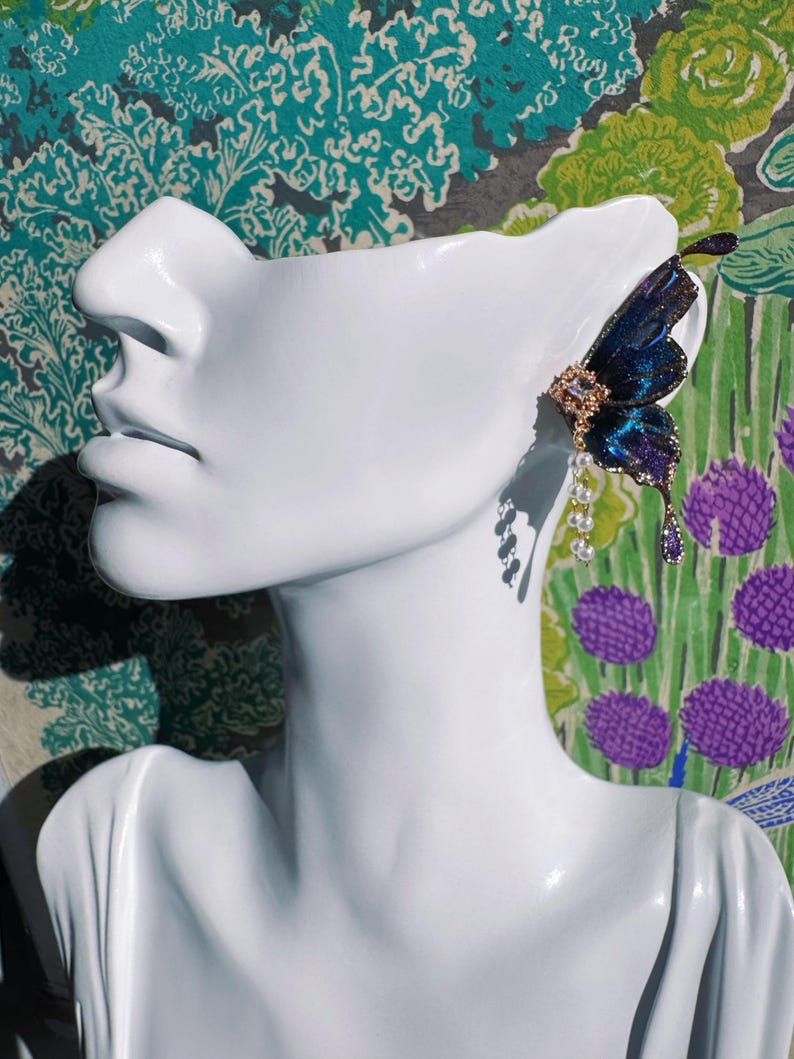 Puede incluir: Un pendiente de oreja con forma de mariposa en tonos azules y morados, adornado con detalles dorados y colgantes de perlas y gemas moradas. El pendiente se exhibe en una cabeza de maniqu&iacute; blanca sobre un fondo floral colorido.