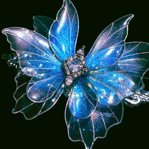 May include: A butterfly-shaped hair accessory featuring translucent, iridescent blue wings with a glittery finish and silver-toned wire edges. The center of the butterfly is adorned with a square-cut clear crystal surrounded by smaller clear stones.