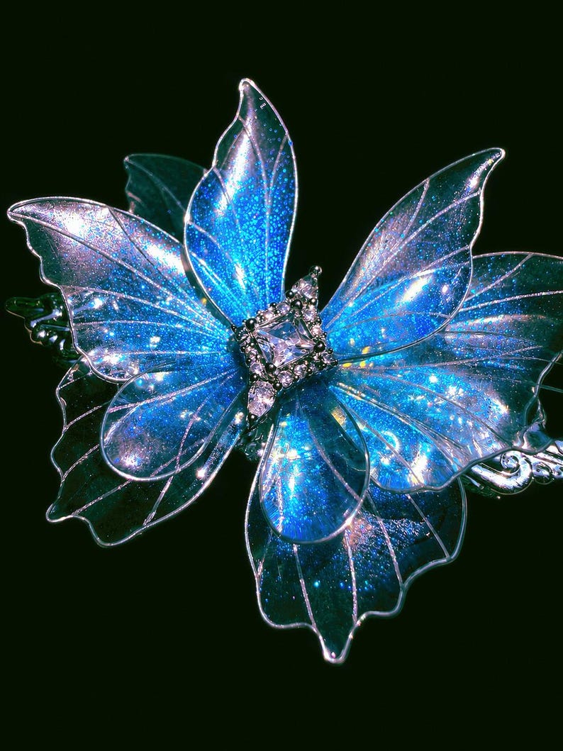 May include: A butterfly-shaped hair accessory featuring translucent, iridescent blue wings with a glittery finish and silver-toned wire edges. The center of the butterfly is adorned with a square-cut clear crystal surrounded by smaller clear stones.