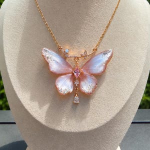 May include: A gold-toned necklace featuring a large butterfly pendant with translucent pink and white wings accented by silver glitter. The center of the butterfly is set with a pink marquise-cut stone, and a clear teardrop crystal hangs from the bottom.