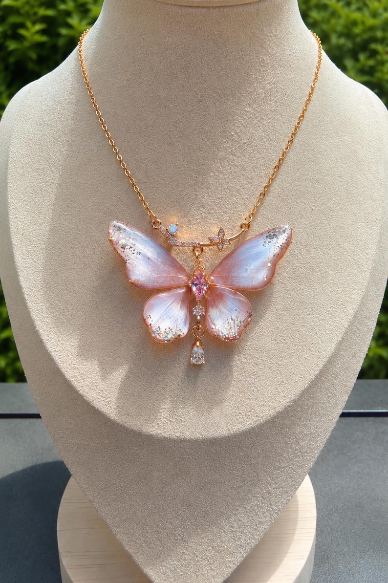 May include: A gold-toned necklace featuring a large butterfly pendant with translucent pink and white wings accented by silver glitter. The center of the butterfly is set with a pink marquise-cut stone, and a clear teardrop crystal hangs from the bottom.