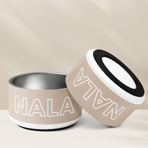 Puede incluir: Dos cuencos para mascotas de color beige con un borde negro y un contorno blanco del nombre "NALA" en cada cuenco.