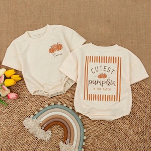 May include: Two cream-colored short-sleeve rompers. The left romper features a small orange pumpkin graphic and the name Thomas. The right romper displays a large orange striped frame with the text Cutest pumpkin in the patch.