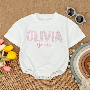May include: White baby romper with the name "Olivia Grace" in pink floral letters. The romper has short sleeves and snap closures at the bottom. Sunglasses and a rainbow decoration are also visible.