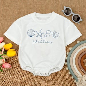 May include: White baby romper with a crew neck and short sleeves. The romper features a blue seashell and starfish design with the name "William" in script. Includes snap closures at the bottom.