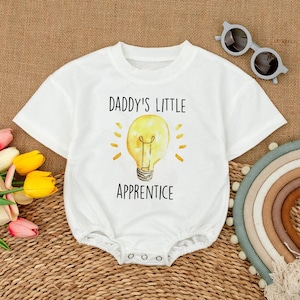 May include: White baby onesie with the text "DADDY'S LITTLE APPRENTICE" above a yellow lightbulb graphic. The onesie has short sleeves and a snap closure. The background is a woven texture.
