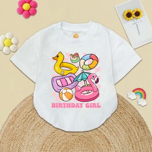 May include: White t-shirt with a colorful graphic of pool floats, including a flamingo, a duck, a raft, and a ring. The text "Birthday Girl" is printed below the graphic.