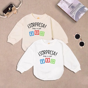 May include: Two white long-sleeved shirts with the text "¡Sorpresa! Vas a ser Tío" in black lettering with colorful block letters. The shirts are on a light brown surface.