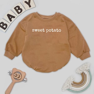 May include: A brown baby onesie with the words "sweet potato" printed in white. The onesie has long sleeves and a round neckline. Baby blocks spelling "BABY" are in the upper left corner. A wooden rattle and a rainbow are also in the image.
