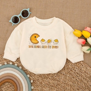 Op de afbeelding: Crèmekleurige babyromper met een afbeelding van een taartvormig personage en drie gevulde eieren. De tekst "YOU KNOW WHY I'M HERE" staat eronder gedrukt. Een lichtblauwe zonnebril en een boeket bloemen zijn ook zichtbaar.