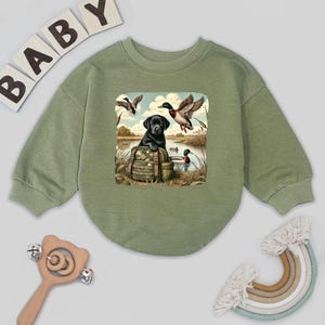 May include: Olive green baby sweatshirt with a graphic of a black Labrador puppy sitting on a backpack, with ducks flying and swimming in a lake. The word "BABY" is spelled out in wooden letters above the sweatshirt.